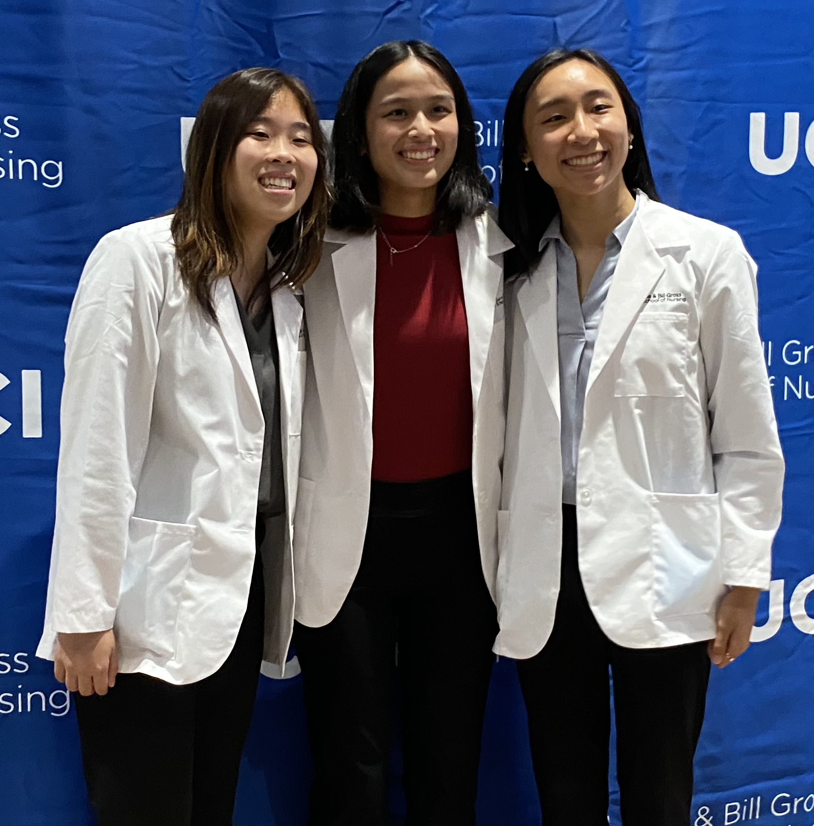three uc irvine school of nursing students class of 2023 in wearing white coats front of blue and white step and repeat