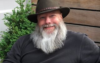 uci department of logic and philosophy professor kyle stanford with white beard, brown shirt, brown hat, smiling