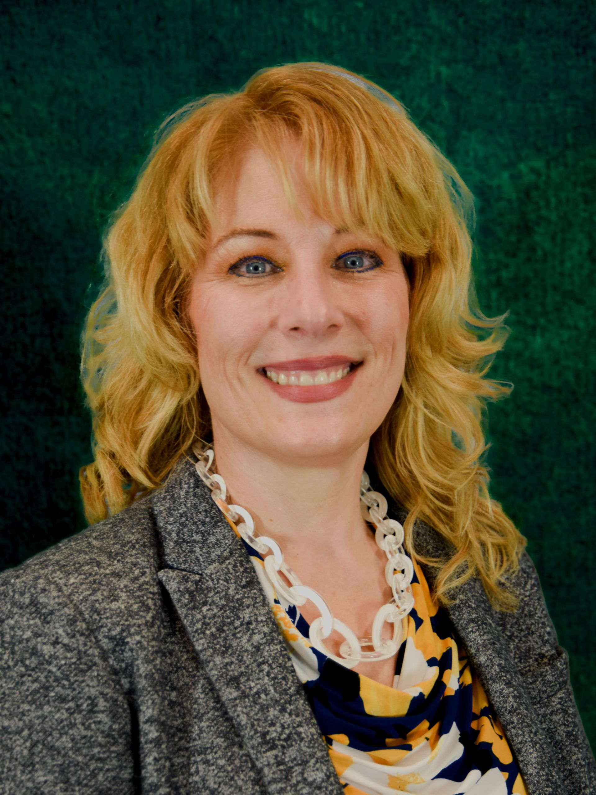 studio image of tiffany nielsen wearing black shirt, gray jacket in front of blue backdrop